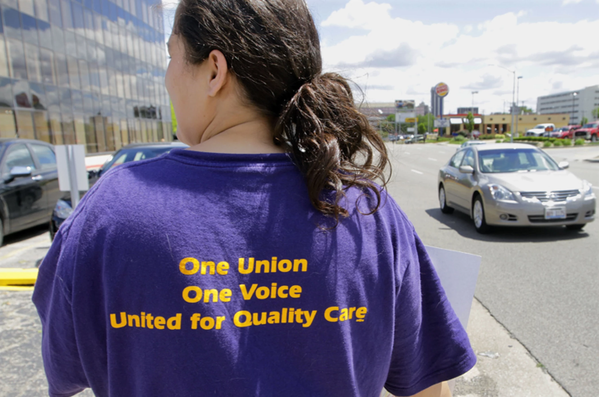 Woman with a long ponytail stands in a parking lot. Her shirt is SEIU branding (purple and gold) and reads "One Union/ One Voice/ United for Quality Care"