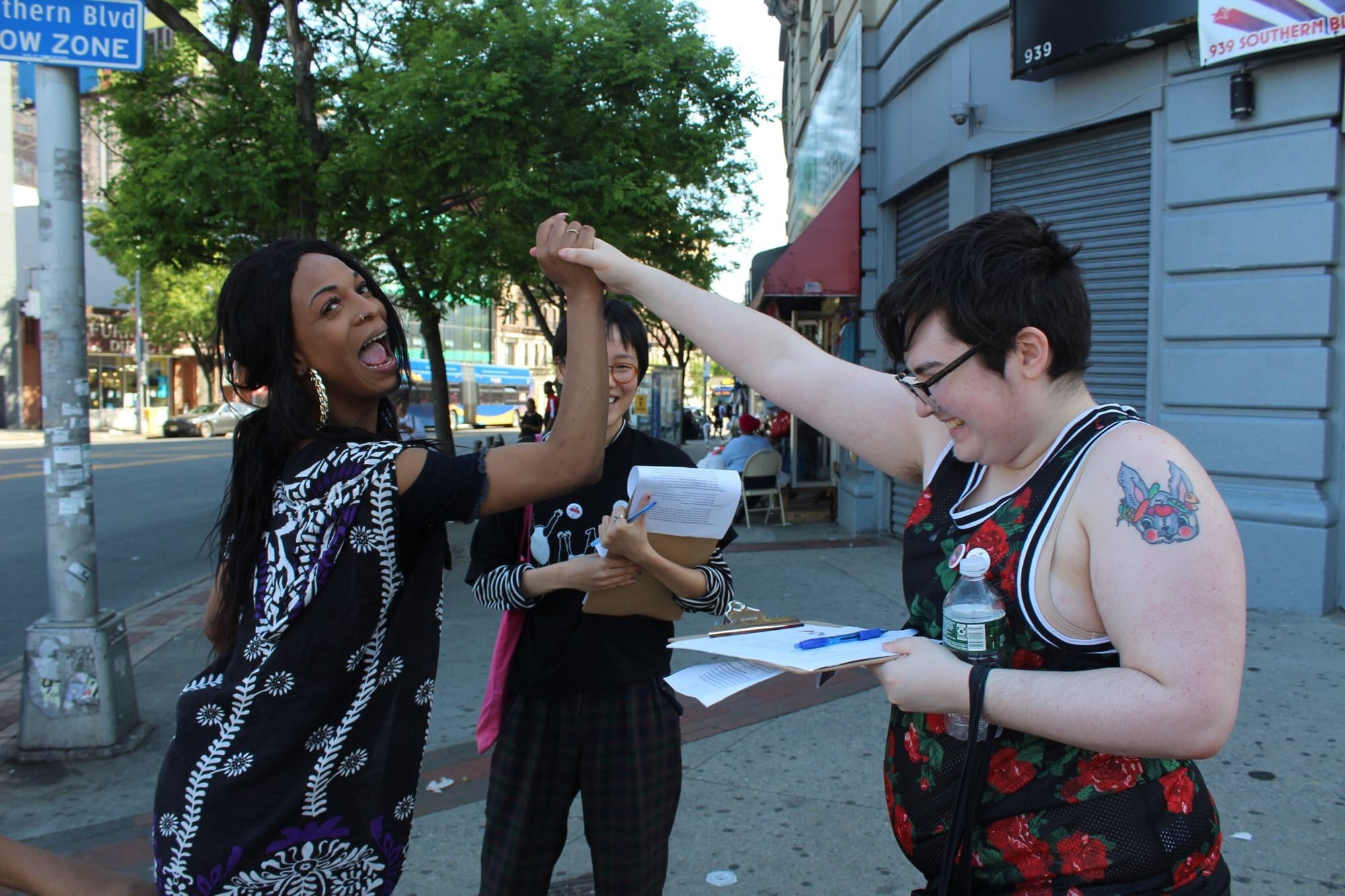 Noah, a white trans man, is shaking hands in victory with Candii, a black trans woman. There is one other canvasser in the background, all holding clipboards.