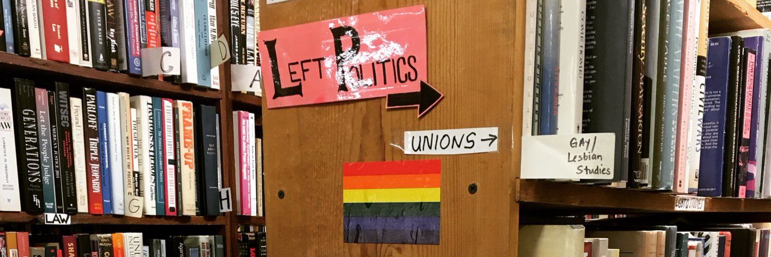 Bookstore shelves with signage reading "left politics" "unions" "gay/lesbian studies" and a rainbow flag.